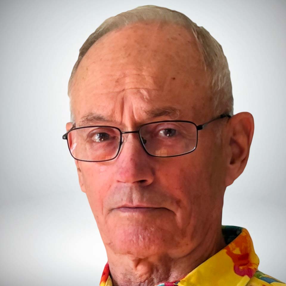 Elderly man with short gray hair and glasses, wearing a colorful shirt, looking directly into the camera against a plain background.