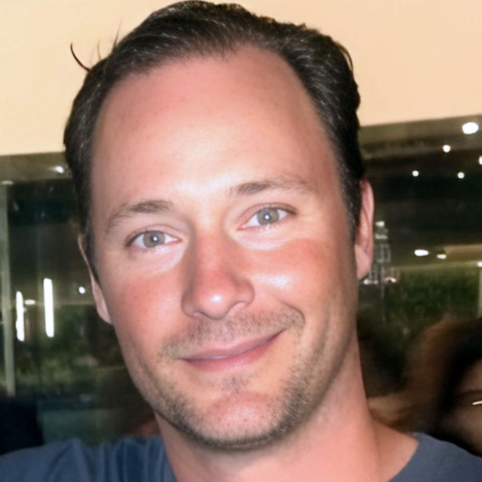 A man with short brown hair and light facial hair smiles at the camera. He is wearing a blue shirt. The background appears to be indoors with some lights and a window reflecting outside scenery.