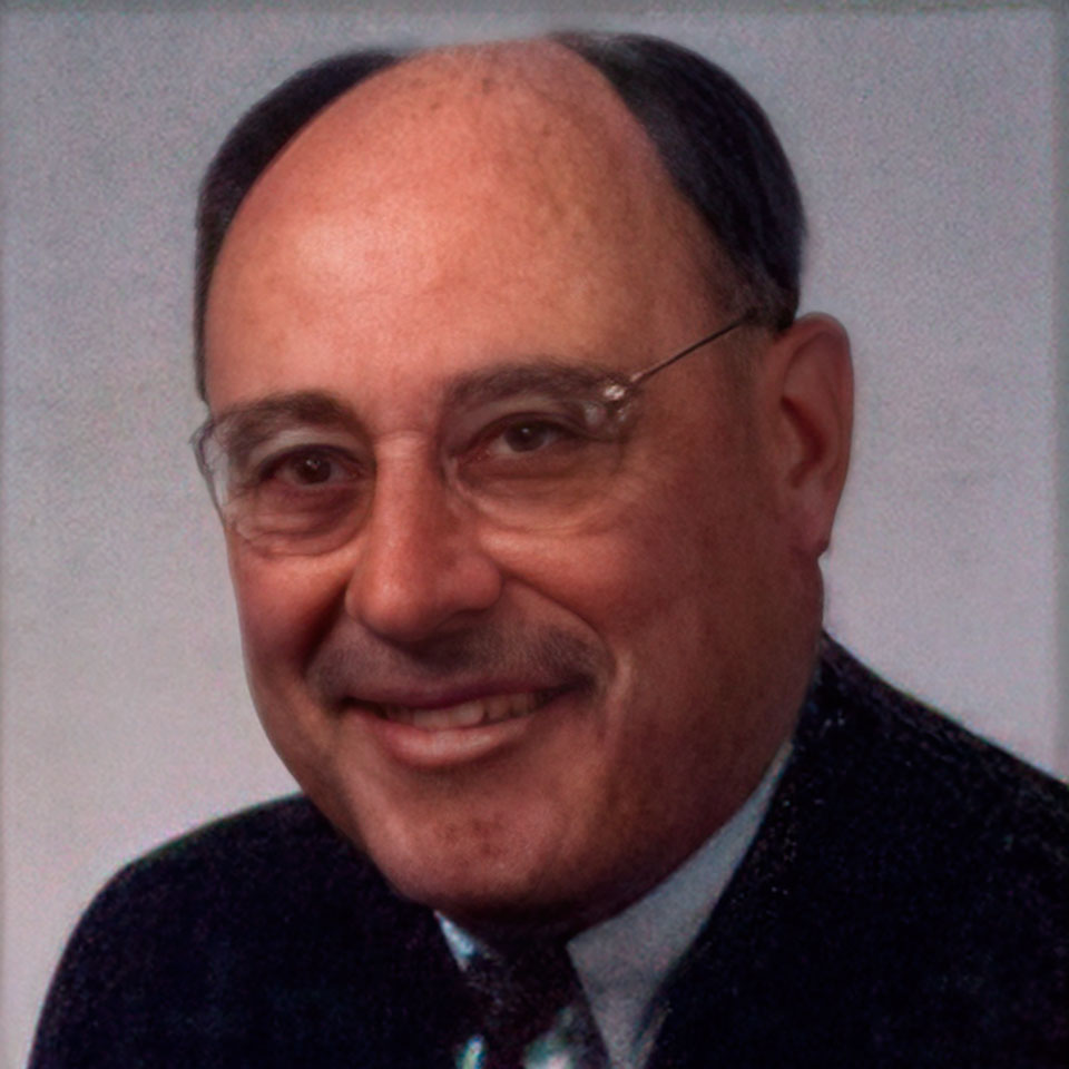 A portrait of an older man with glasses and a bald top, wearing a suit and tie. He is smiling and has a mustache. The background is a plain, light gray color.