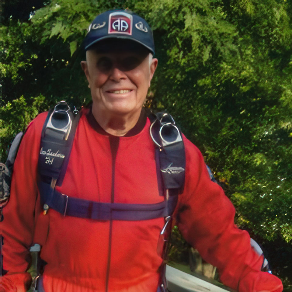 An elderly person wearing a red parachute jumpsuit and a cap stands outdoors. They are smiling, with trees in the background. The parachute straps are visible on their shoulders.