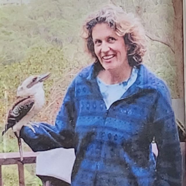 A woman with curly hair, wearing a blue jacket, smiles while standing on a balcony with a kookaburra perched on her hand. The background shows lush green trees.