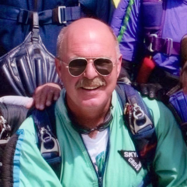 A man wearing sunglasses and a light green jumpsuit smiles at the camera. He has a mustache and is in front of a group of people wearing parachute gear.