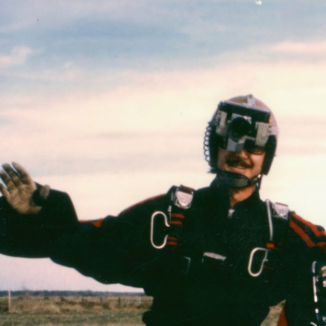 A person in a black and red skydiving suit and helmet waves at the camera. The helmet has a mounted camera. The sky is slightly cloudy, and there is a grassy landscape in the background.