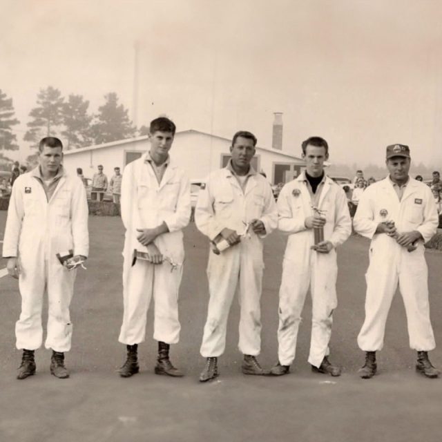 Five men in white jumpsuits and boots stand in a row on a paved area. A single-story building and trees are visible in the background. They all hold objects in their hands and appear to be posed for the photo.