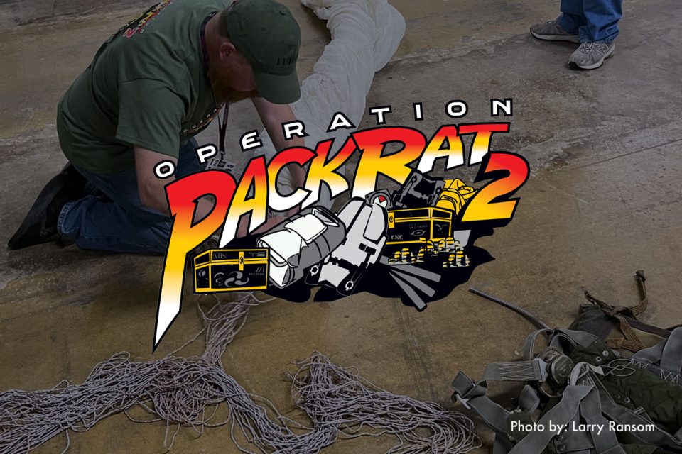 A person in a green shirt works on a bundle of ropes on the floor. The text "Operation Packrat 2" with cartoon vehicles and items is overlaid. Photo credit: Larry Ransom.