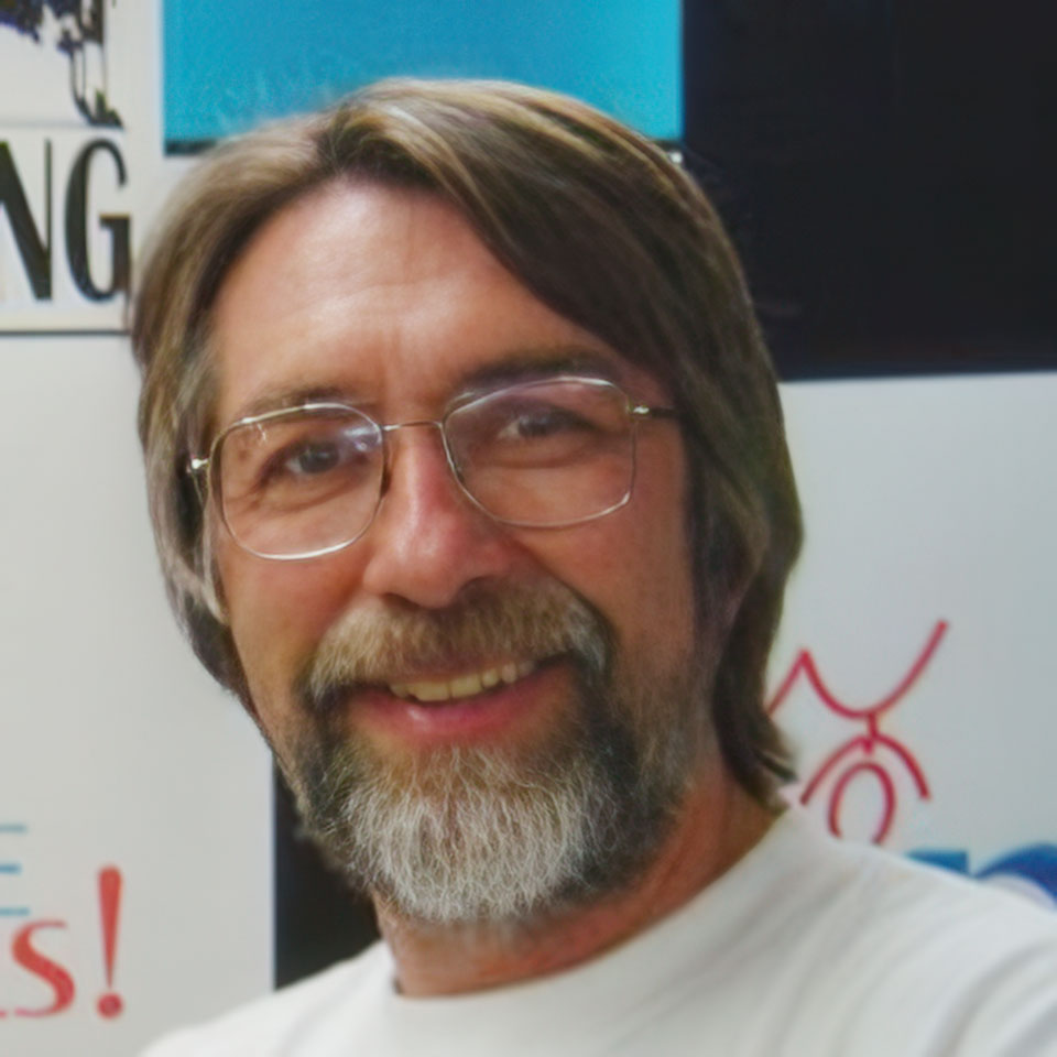 A man with glasses, a beard, and long brown hair is smiling. He is wearing a white shirt and standing in front of a colorful background with text and graphics.