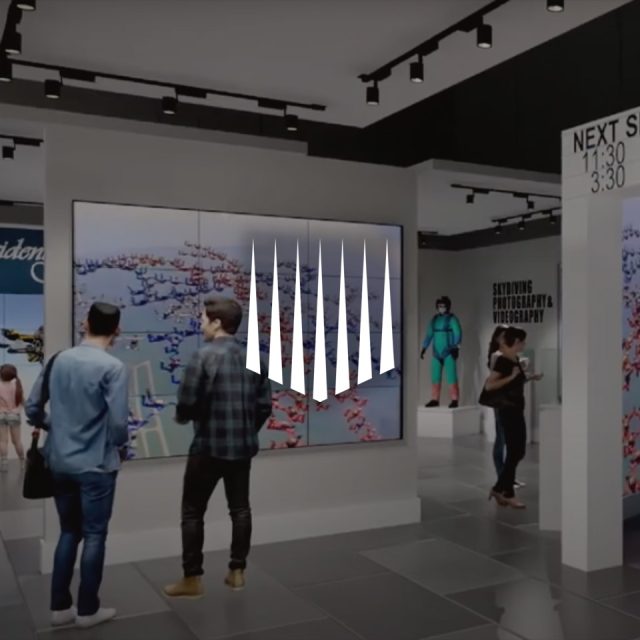 People stand in a modern museum or gallery space, observing a large screen displaying digital artwork. The environment is sleek and contemporary, with minimalistic design and overhead lighting. Exhibition schedule signs are visible.