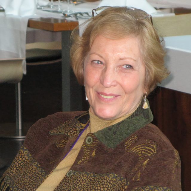 A smiling woman with short, light brown hair is sitting indoors. She is wearing a patterned brown jacket over a beige top. In the background, there are tables set with white tablecloths.