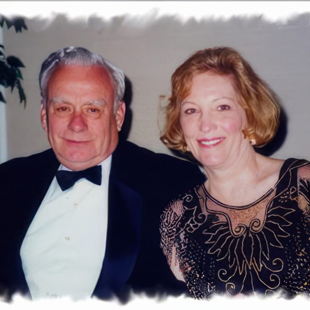 A man in a tuxedo and a woman in a black dress with gold embroidery smile while sitting together indoors. The background is softly blurred.