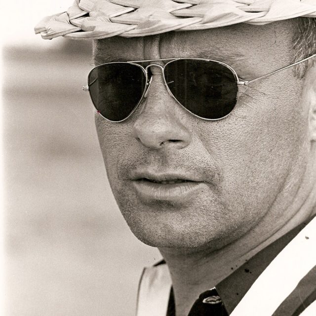 A black and white photo of a man wearing aviator sunglasses and a woven straw hat. His expression is serious, with a hint of stubble on his face. The image focuses closely on his head and shoulders.