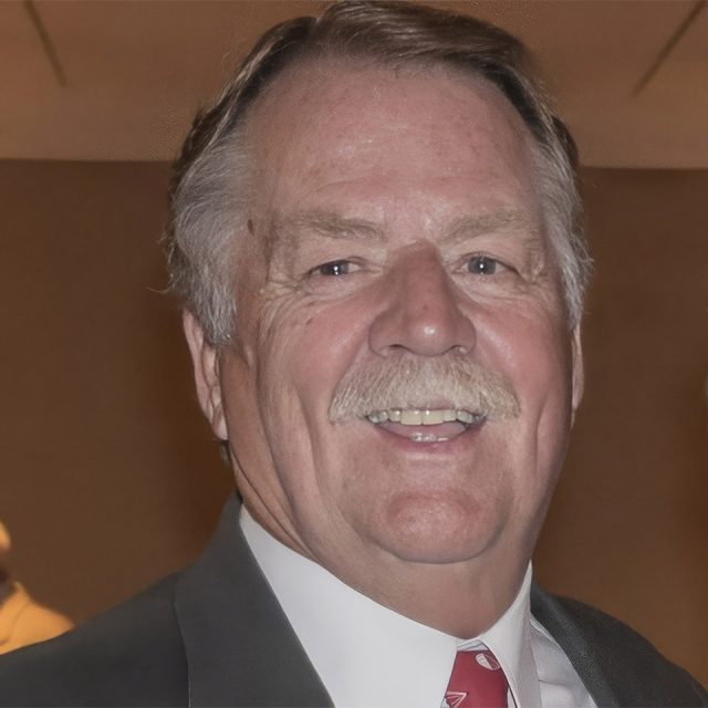 A mature man with gray hair and a mustache is smiling. He is wearing a suit and red tie. The background is a blurred indoor setting.