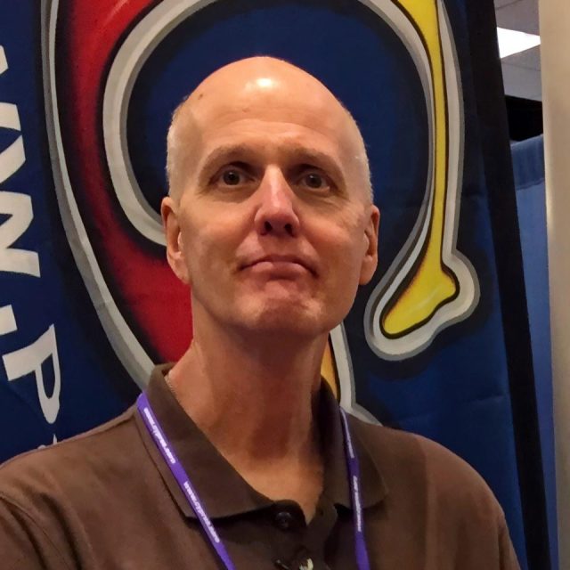 A bald man wearing a brown shirt and a purple lanyard stands in front of a colorful backdrop with abstract designs. He has a neutral expression and the background features a combination of blue, red, and yellow shapes.