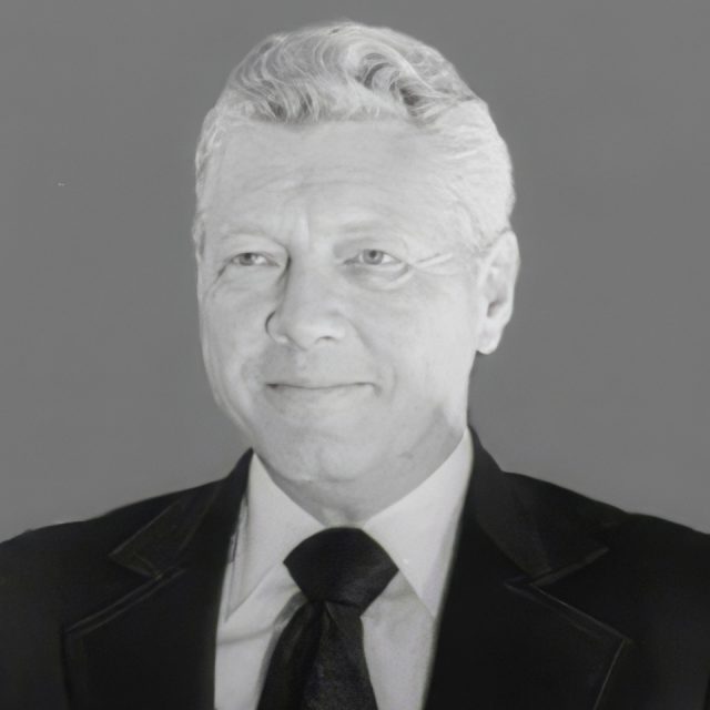 A grayscale portrait of an elderly man with gray hair, wearing a dark suit and tie. He has a slight smile and is looking slightly to the side. The background is plain.