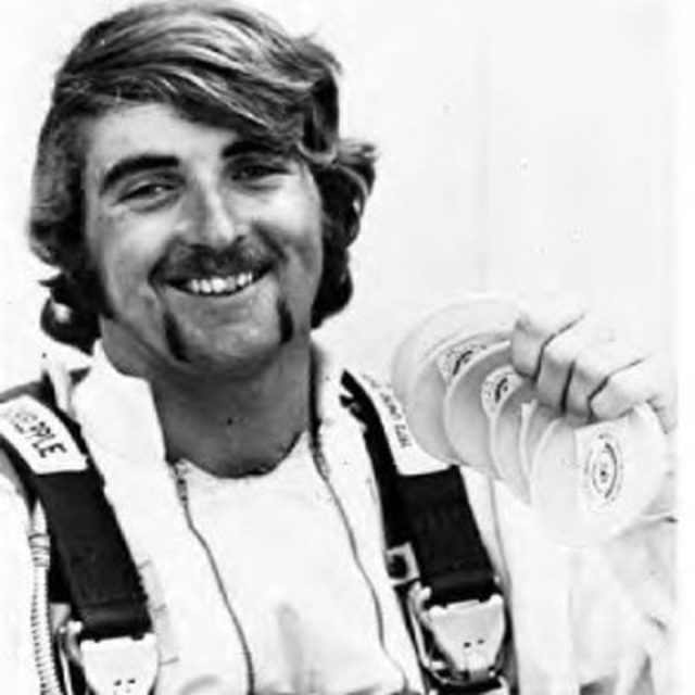 A person smiling with a thick mustache and long sideburns, wearing a white uniform and harness, holding several frisbees. The background is plain white.