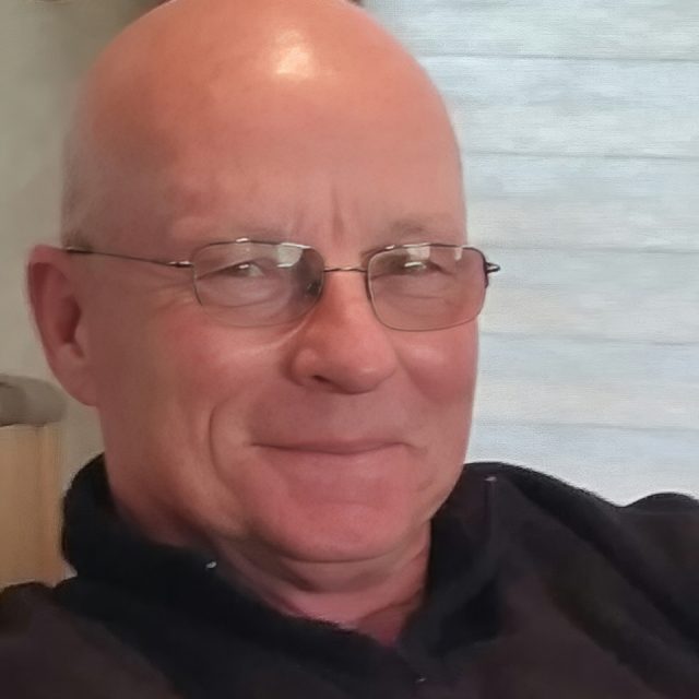 Bald man wearing glasses, smiling, and dressed in a dark shirt, seated indoors with a light-colored background.