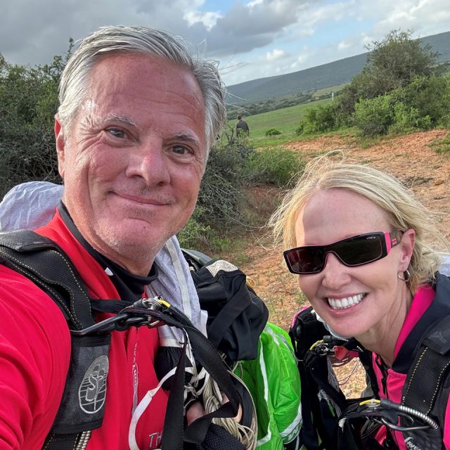 A man and woman smiling outdoors, each wearing red attire and harnesses. They stand in a grassy landscape with hills in the background. The sky is partly cloudy. The woman is wearing sunglasses. They appear ready for an outdoor activity.