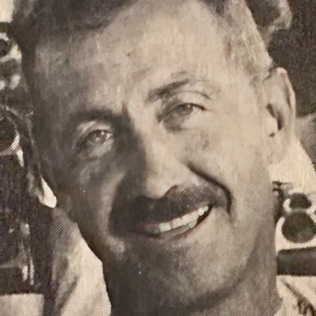 A grayscale portrait of a smiling man with short hair and a mustache. The background is blurred, emphasizing the man's face.