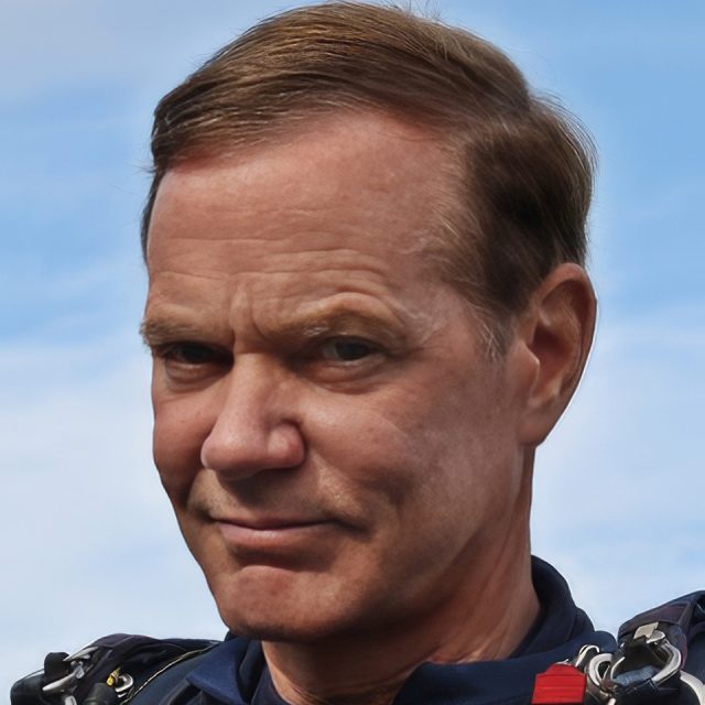 A person with short hair wearing a blue jumpsuit and a parachute harness is outdoors against a backdrop of a clear blue sky. The expression is neutral, and they are looking directly at the camera.