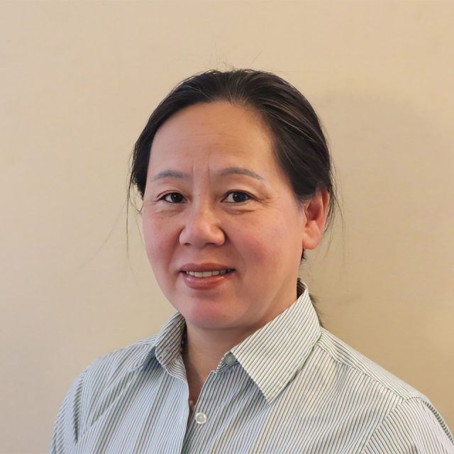 A person with dark hair tied back is wearing a light striped shirt and smiling slightly at the camera against a plain beige background.