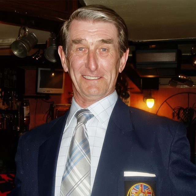 An older man in a blue blazer with a crest on the pocket, wearing a checked shirt and plaid tie, smiles in a warmly lit indoor setting. He has short gray hair and the background features a bar area with hanging lights.