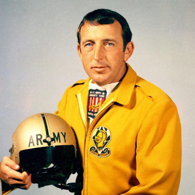 A man in a yellow jacket holds a helmet labeled "ARMY." The jacket has various patches, including one saying "Golden Knights." He has short dark hair and a serious expression. The background is a plain light color.