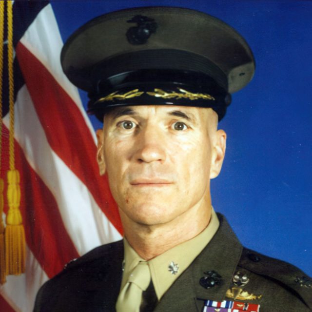 A uniformed military officer stands in front of an American flag. He wears a brown service cap and jacket adorned with various medals and insignia. The background is a solid blue.