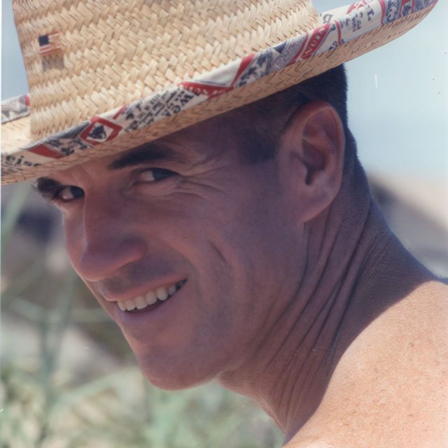 A person wearing a straw hat with a patterned band smiles at the camera. They are outdoors with a blurred background that suggests a sunny setting.