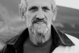 A grayscale portrait of an older man with a beard and short hair. He is wearing a jacket over a T-shirt, looking directly at the camera. The background is blurred, focusing on his face.