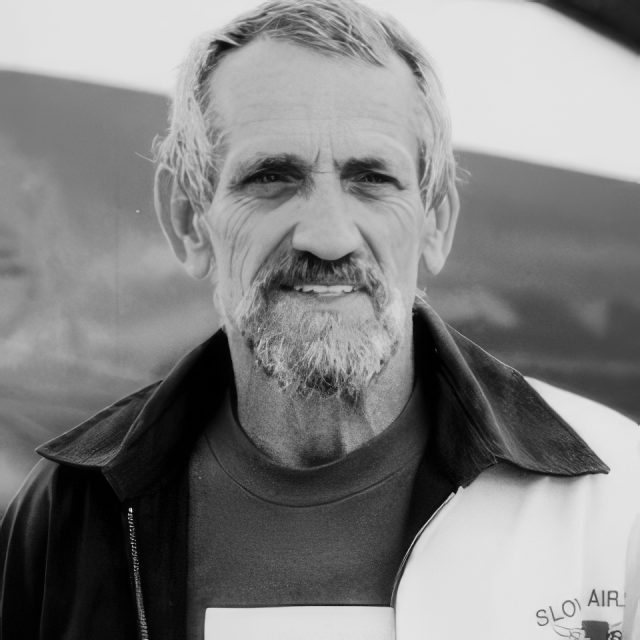 A grayscale portrait of an older man with a beard and short hair. He is wearing a jacket over a T-shirt, looking directly at the camera. The background is blurred, focusing on his face.