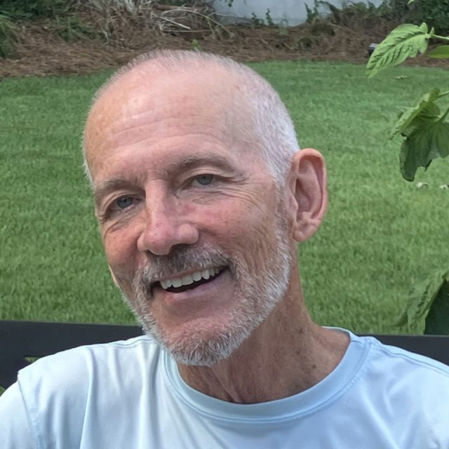 A smiling older man with a shaved head and gray beard is sitting outdoors. He's wearing a light blue shirt, and the background features green grass and some plants.