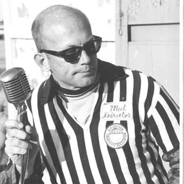 A man with sunglasses and a bald head wearing a referee shirt holds a vintage microphone. A badge on his shirt reads "Meet Director" and "Eastern Association." He appears to be outdoors.