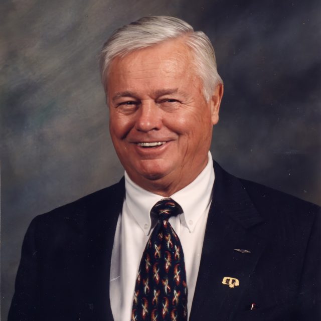 An older man with white hair is wearing a dark suit, a white shirt, and a patterned tie. He is smiling and posed against a dark, neutral background.