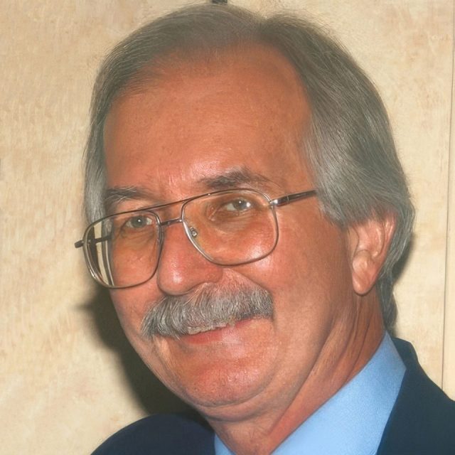 A man with gray hair and a mustache is smiling. He is wearing glasses and a blue shirt with a dark suit. The background is a neutral beige color.