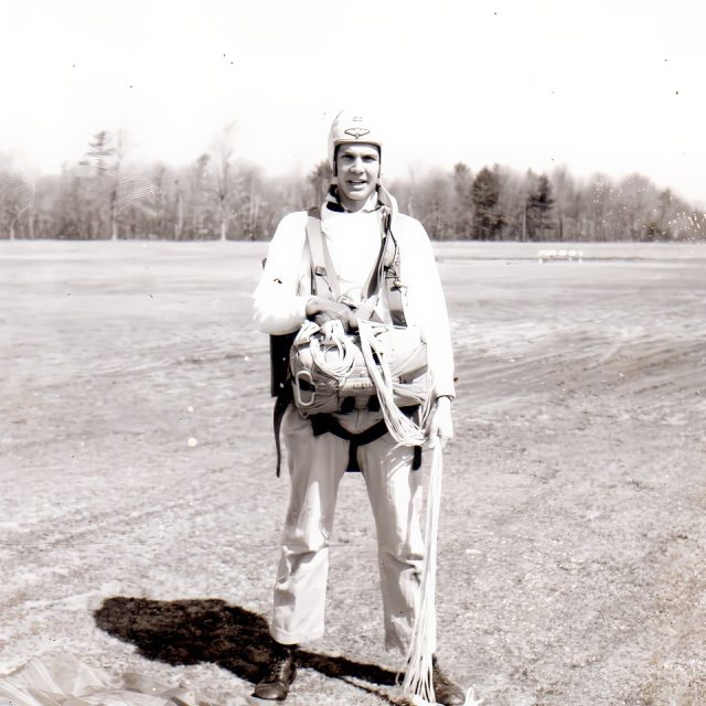 A person stands outdoors on grass, wearing a parachute harness and holding parachute cords. They wear a helmet and appear ready for skydiving. Trees can be seen in the background under a clear sky. The image is in black and white.