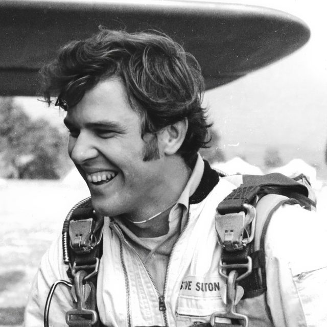 A smiling man in a parachute harness and jumpsuit stands outdoors. The image is black and white, with a blurred background featuring trees and part of an aircraft wing.