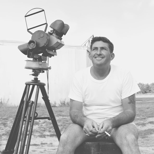 A man in a white t-shirt and shorts, sitting on a wooden block outdoors, smiling while holding a cigarette. Next to him is a large telescope mounted on a tripod. The background shows a clear sky and a building.