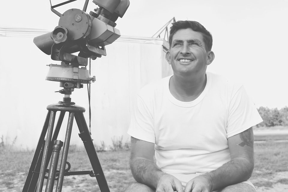 A man in a white t-shirt and shorts, sitting on a wooden block outdoors, smiling while holding a cigarette. Next to him is a large telescope mounted on a tripod. The background shows a clear sky and a building.