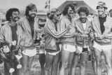 A group of six men in matching athletic outfits, smiling and holding trophies. They stand outdoors on grass, with some wearing sunglasses and hats. One holds a toy monkey. The scene is lively and celebratory.