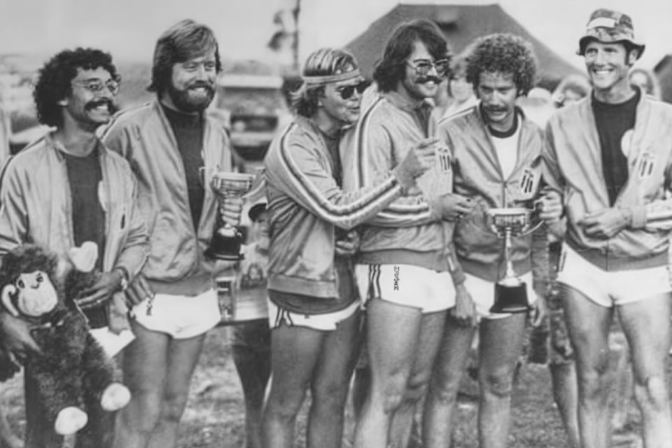 A group of six men in matching athletic outfits, smiling and holding trophies. They stand outdoors on grass, with some wearing sunglasses and hats. One holds a toy monkey. The scene is lively and celebratory.