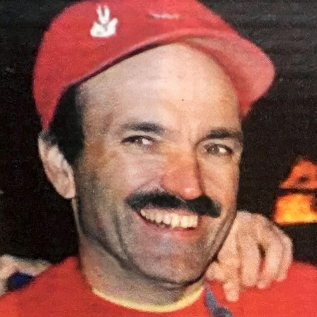 A person wearing a red cap with a logo and a matching red shirt smiles warmly. They have a mustache and are photographed with a slightly blurred background that appears dark.