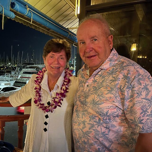 An elderly couple stands together on a balcony at night, overlooking a marina with boats. The woman wears a floral lei and a white outfit, while the man wears a patterned shirt. They are smiling, enjoying the evening.