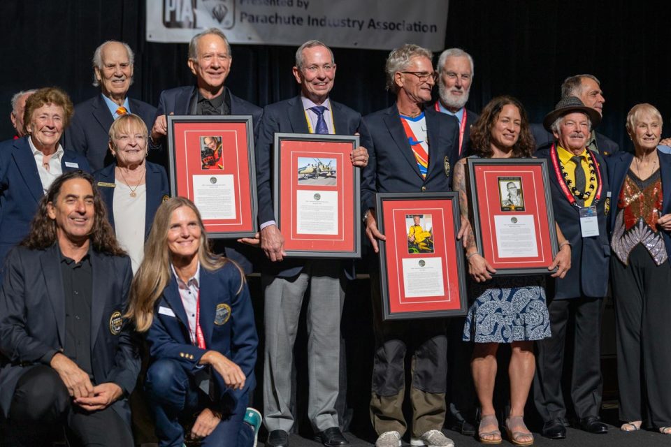 A group of people, including men and women, stand on a stage holding framed certificates. They are smiling and dressed in formal and casual attire. A backdrop with text and logos is partially visible behind them.