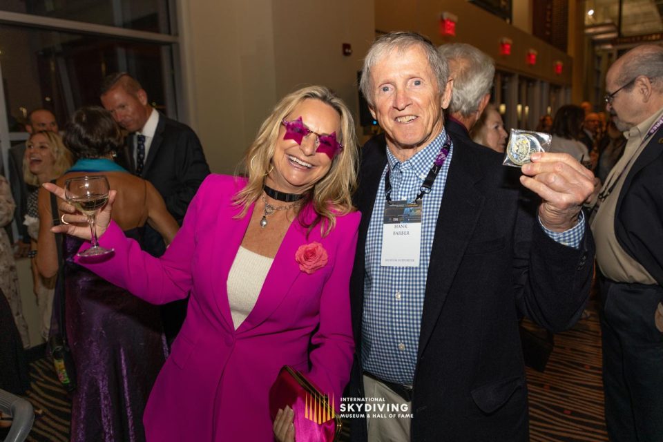 A woman in a pink outfit and star-shaped glasses poses with a man in a suit holding a medal. They are smiling at a formal event with others in the background. A sign reads, "International Skydiving Museum & Hall of Fame" on the carpet.