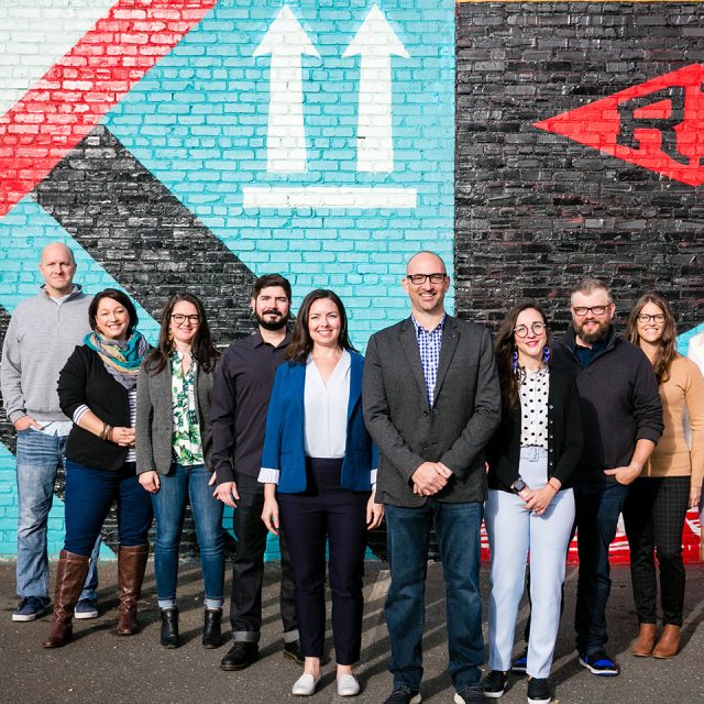 A diverse group of ten people standing in front of a colorful brick wall with geometric designs. They are smiling and casually dressed, with some wearing glasses. The wall features red, black, and turquoise patterns with two white arrows.