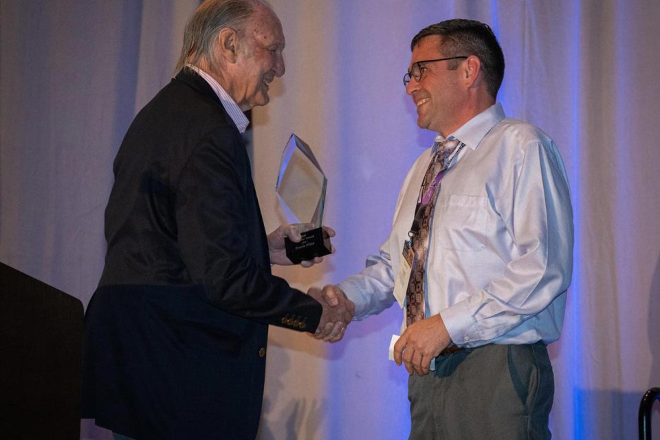 Two men are shaking hands on stage during an award ceremony. One man is holding a clear trophy. Both are smiling and dressed in formal attire. The background features curtains, and the event is related to the International Skydiving Museum.