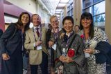 A group of five people, dressed in formal and traditional attire, stand smiling and holding drinks in a conference hallway. They wear attendee badges and appear to be enjoying a social gathering.