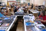 People sitting at tables in a warehouse setting, organizing stacks of aviation magazines and publications. Shelves and boxes are visible in the background, indicating a sorting or packaging activity.