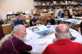 A group of people seated around a table in a busy room with shelves of boxes. They are engaged in conversation, some holding drinks. A whiteboard and various papers are on the table.