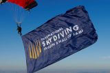 A skydiver descends with an open parachute, holding a large blue flag that reads "International Skydiving Museum & Hall of Fame." The background shows a landscape of fields under a clear blue sky.