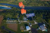 Aerial view of three skydivers with orange parachutes descending over a lush green landscape with a river. Below, a campsite with numerous white trailers and a few buildings is visible.